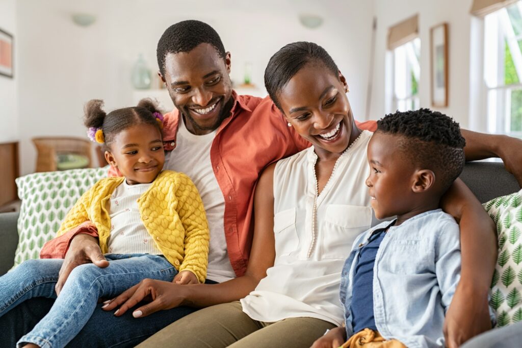 Happy Black family enjoying quality time together at home, with parents engaging with their two children on a cozy couch, emphasizing warmth and family bonding in a comfortable living space.