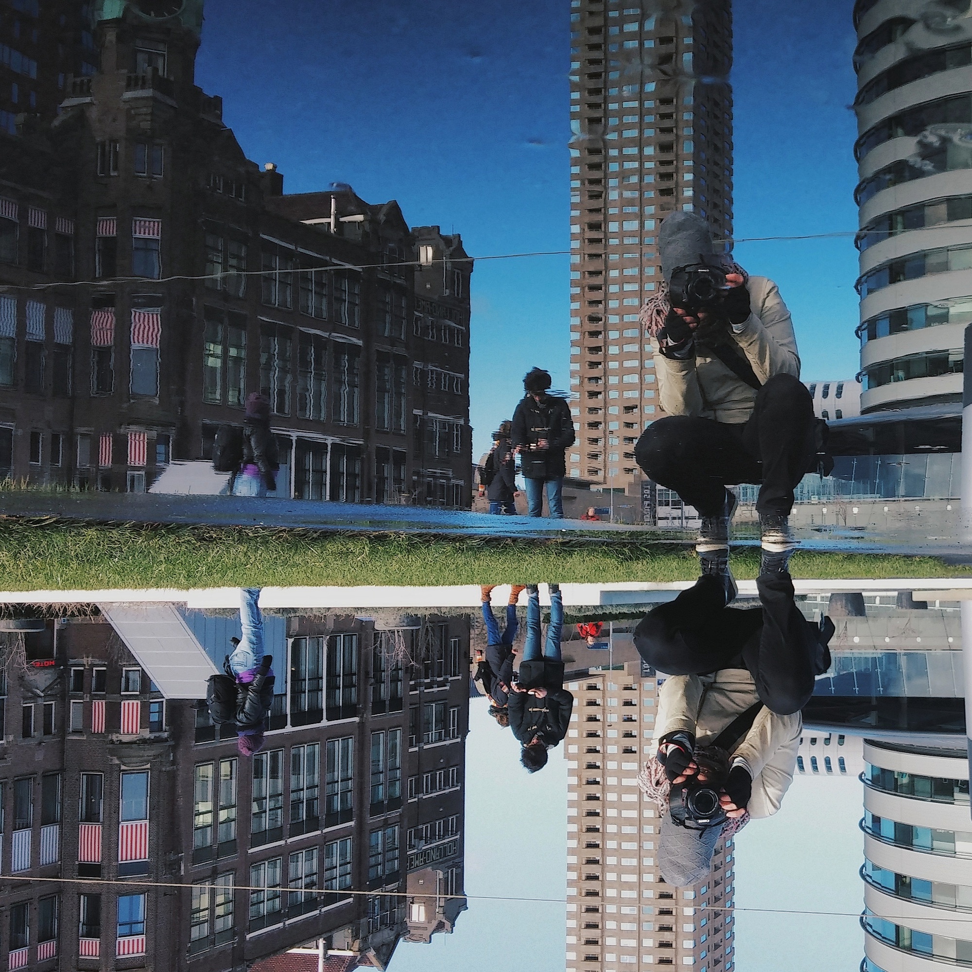 Person taking a photo with a camera, reflecting in a puddle, surrounded by urban architecture and pedestrians.