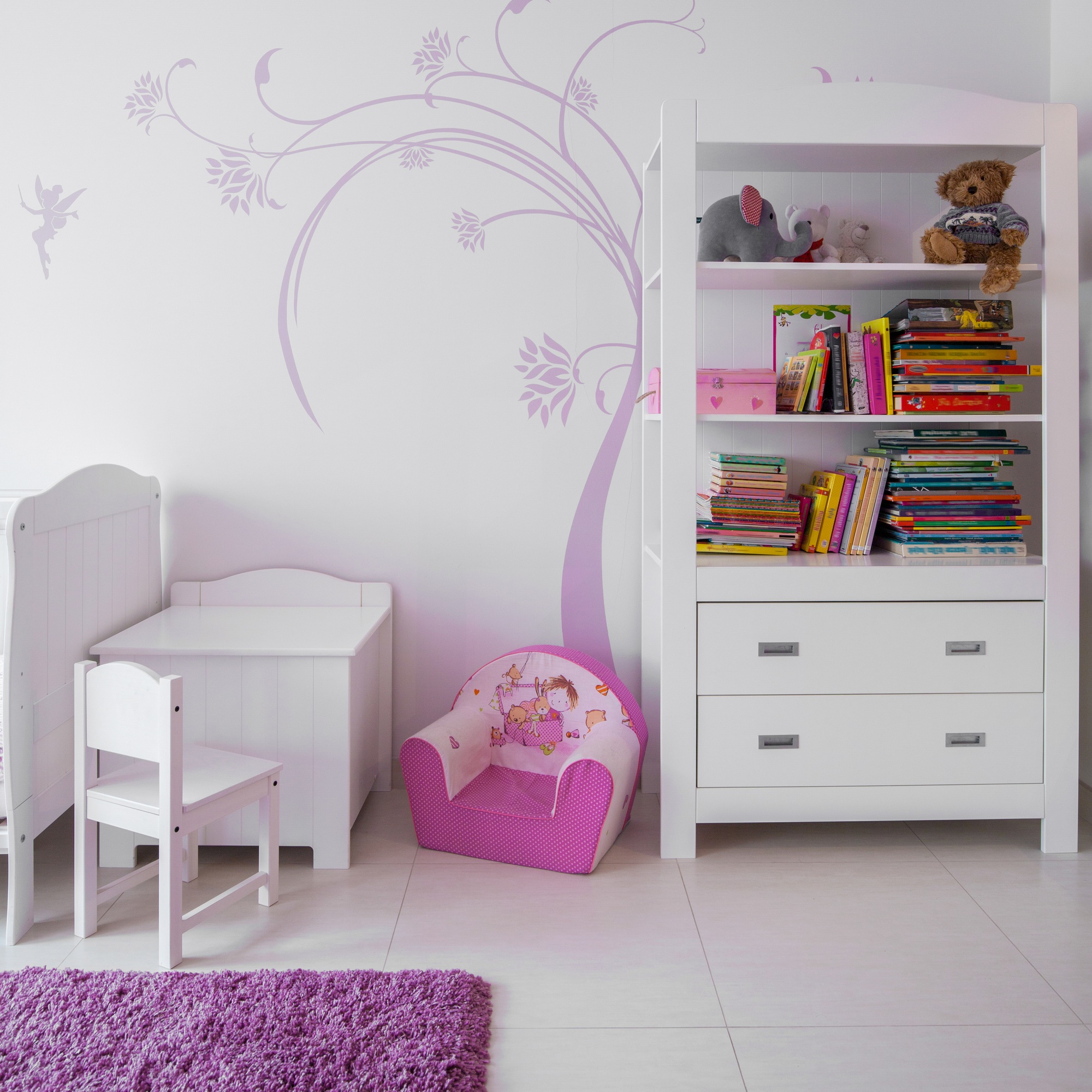 Child's bedroom featuring a white bookshelf filled with colorful books, a small white table and chair, a pink children's armchair, and whimsical wall decor of a tree and fairy.