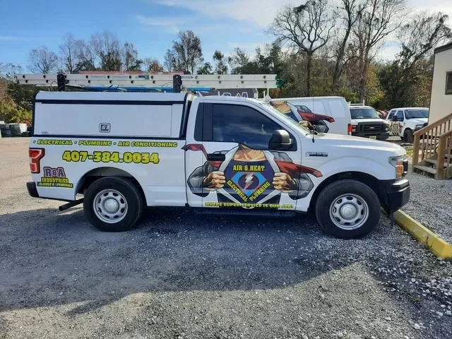 R&A Industries service truck displaying plumbing and air conditioning services, with contact information and a superhero graphic, parked in a service area.