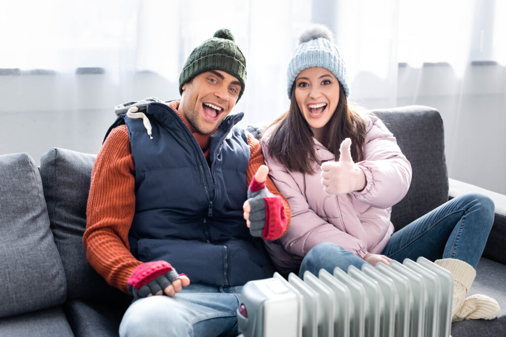 Smiling couple in winter outfits giving thumbs up beside a radiator, highlighting warmth and comfort in home heating.