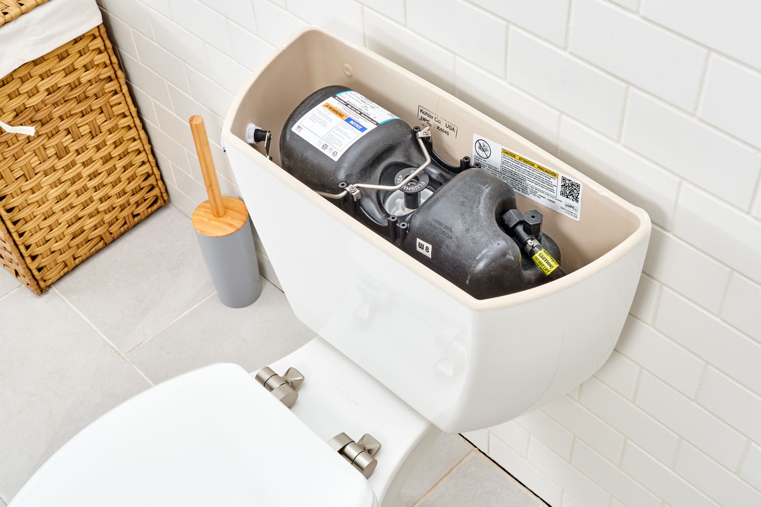 Pressure-assisted toilet tank with internal components visible, showcasing modern plumbing technology, alongside a toilet brush and woven storage basket in a clean bathroom setting.