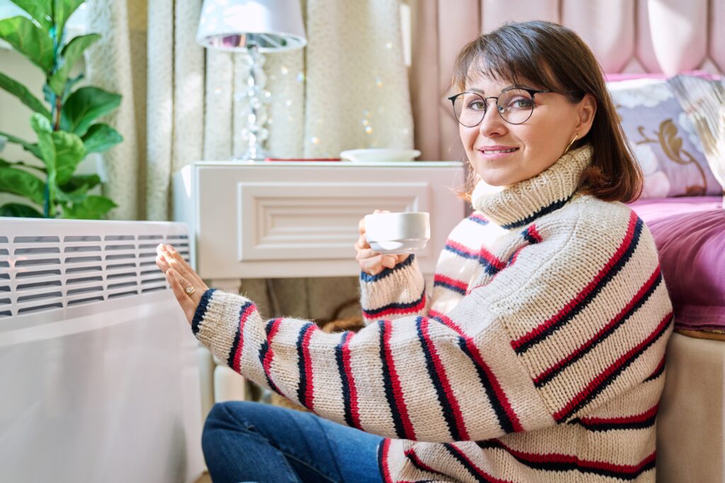 Woman in a cozy striped sweater warming her hands by an electric heater in a well-decorated bedroom, illustrating comfort and warmth related to heat pump services.