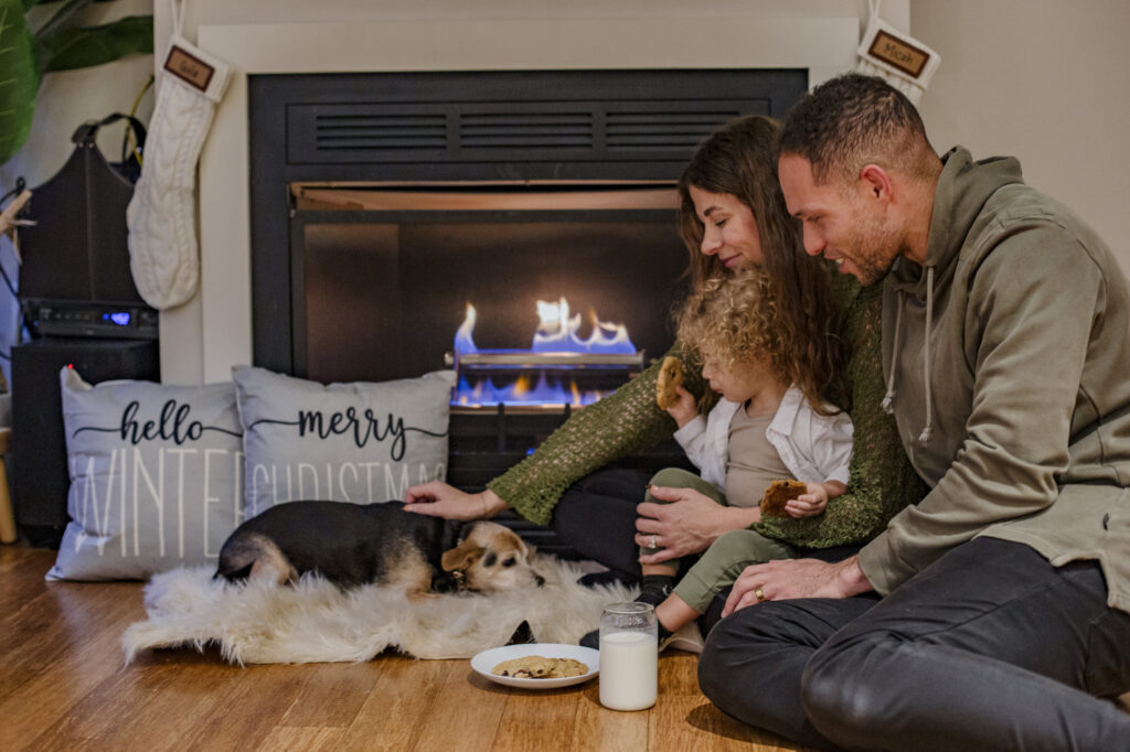Family enjoying cozy moments by a fireplace with a dog, cookies, and milk, emphasizing warmth and comfort during winter.