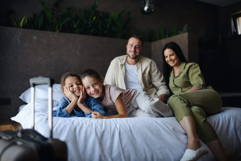 Smiling family relaxes on a bed near luggage, showcasing a comfortable travel experience in Oviedo, FL. Consider EV Charger options nearby.